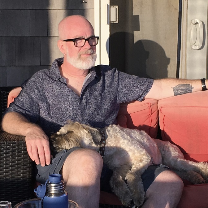 A bald, bearded, older man in casual summer wear sits on patio furniture in bright sunlight with a soft-coated wheaten terrier asleep across his lap.
